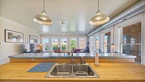 A modern kitchen with a large island, stainless steel sink, pendant lighting, and a view of the city through large windows. - Serendipity House: Historic Downtown Livingston Loft (Livingston)