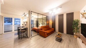 A modern living room with a sofa, a dining table with chairs, a small table with a bottle and glasses, and a large window with a view of buildings. - Urban Chic 2 Luxury Suite Apartments (Roma)