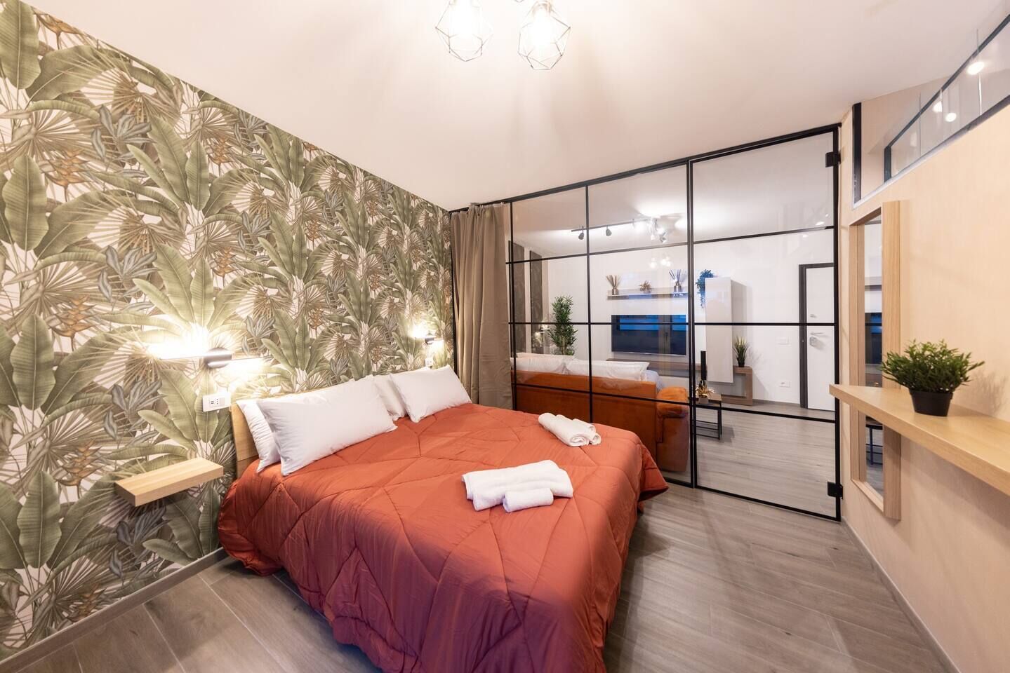 A modern bedroom with a large bed, a sliding glass door leading to a bathroom, and a small plant on a wooden shelf.