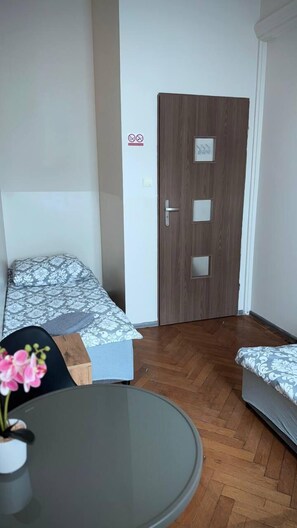A small, compact room with a wooden door, a bed with a patterned bedspread, a small table with a vase of flowers, and a round glass table. - FiveHostel (Kraków)
