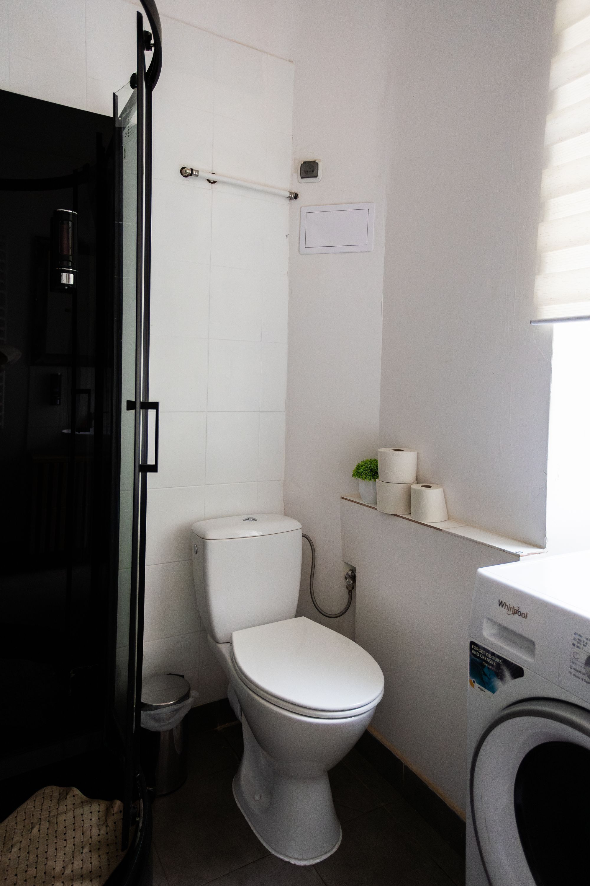 Et lille badeværelse med toilet, en lille skraldespand, bruseområde og en vaskemaskine.