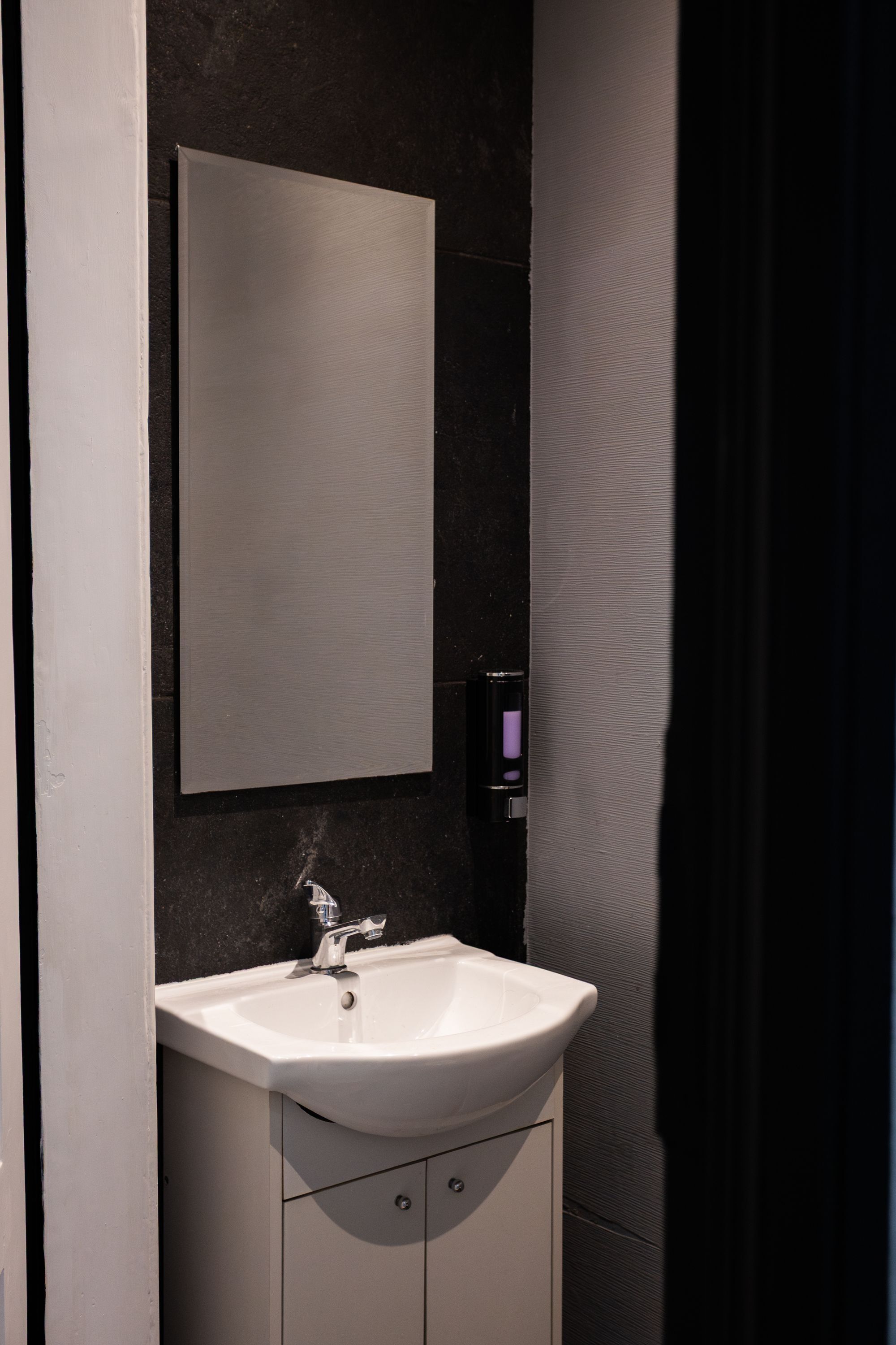 Un baño con lavamanos blanco, espejo y pared oscura con textura.