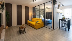 A modern living room with a yellow sofa, a dining area with a table and chairs, and a large sliding glass door leading to a bedroom with a bed and a view of the outdoors. - Urban Chic Luxury Suite Apartments (Roma)
