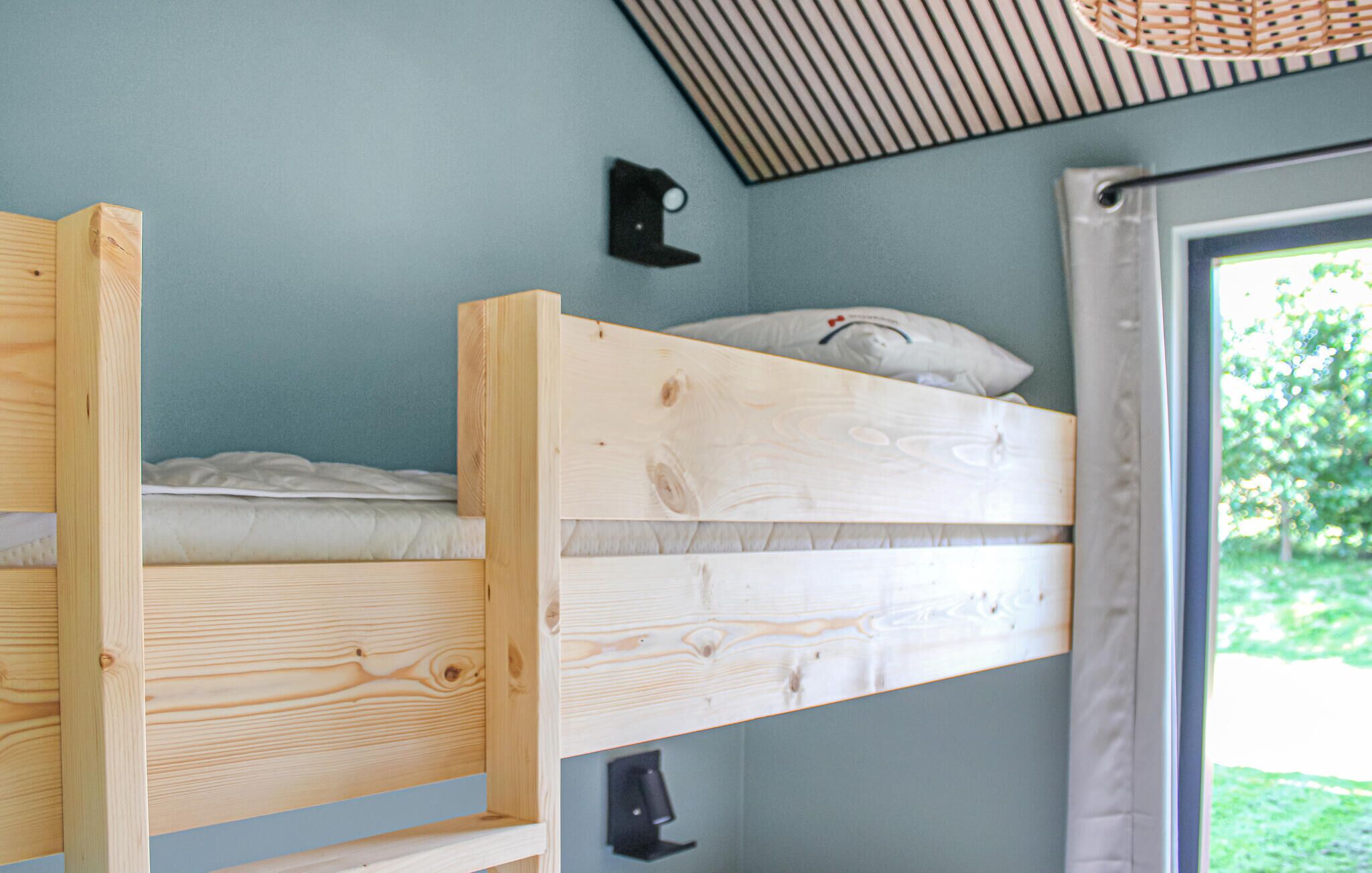 Un lit superposé avec une tête de lit et un oreiller, une petite étagère et une fenêtre donnant sur la verdure.