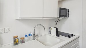Een moderne keuken met een wit aanrecht, een roestvrijstalen spoelbak, een magnetron en een koffieapparaat. Op het aanrecht staan potten met keukengerei en waterflessen.