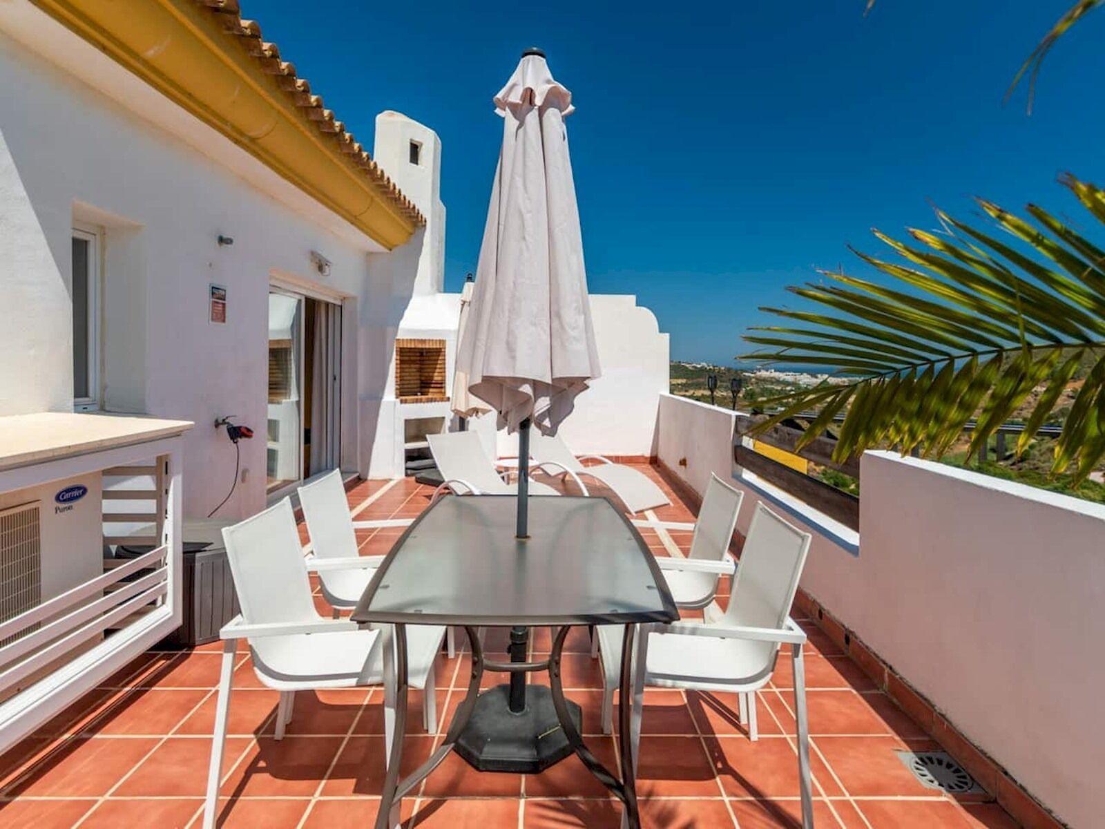 En terrasse med et bord og stole, en hvid parasol og udsigt til havet.