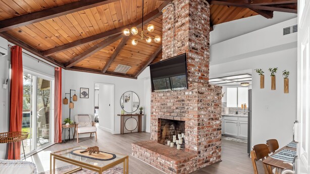 A spacious living area with a brick fireplace, wooden ceiling beams, and a modern kitchen in the background.