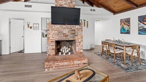 A spacious living room with a brick fireplace, wooden ceiling beams, a flat-screen TV, and a dining area with a table and chairs.