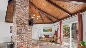 A cozy living room with a brick fireplace, wooden ceiling beams, a sofa, and a large sliding glass door leading to an outdoor area.