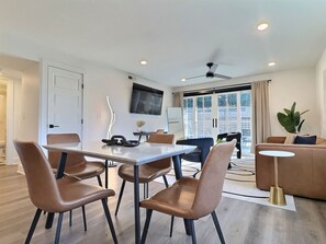 A modern living room with a dining area, a sofa, a TV mounted on the wall, and a large window with curtains. - Upscale suite w/ pool & extra large balcony (Birmingham)