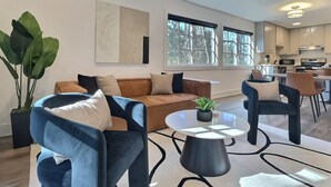A modern living room with a brown sofa, blue armchairs, and a round coffee table. There is a large window providing natural light and a view of the outdoors. - Upscale suite w/ pool & extra large balcony (Birmingham)