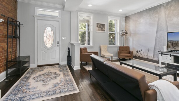 A spacious living room with a large black leather sofa, a glass coffee table, and a patterned rug. There are two armchairs, a fireplace, and a large window with blinds. - 3 DC Homes | Sleeps 30 | Group Getaway (Washington)