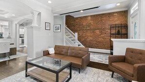 A modern living room with a brown leather sofa, a glass coffee table, and a brick wall in the background. - 3 DC Homes | Sleeps 30 | Group Getaway (Washington)