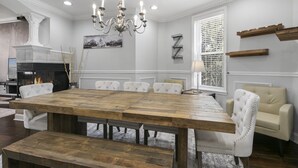 A dining room with a large wooden table, white upholstered chairs, a chandelier, a fireplace, and a window with blinds. - 3 DC Homes | Sleeps 30 | Group Getaway (Washington)