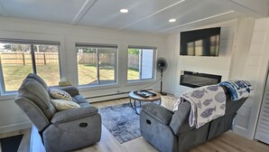 Una sala de estar moderna con dos sofás grises, una pequeña mesa de centro y un televisor montado en la pared. Amplias ventanas ofrecen vistas al patio trasero.