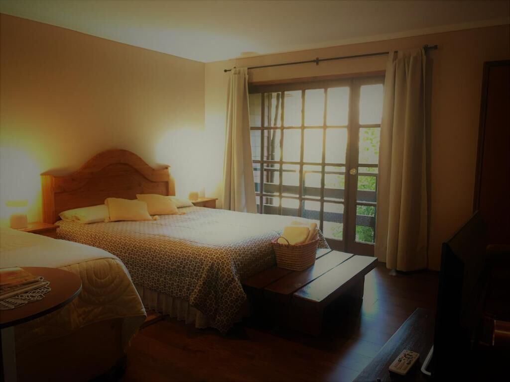 A hotel room with a large bed, a bedside table, a small table, a TV, and a window with curtains.