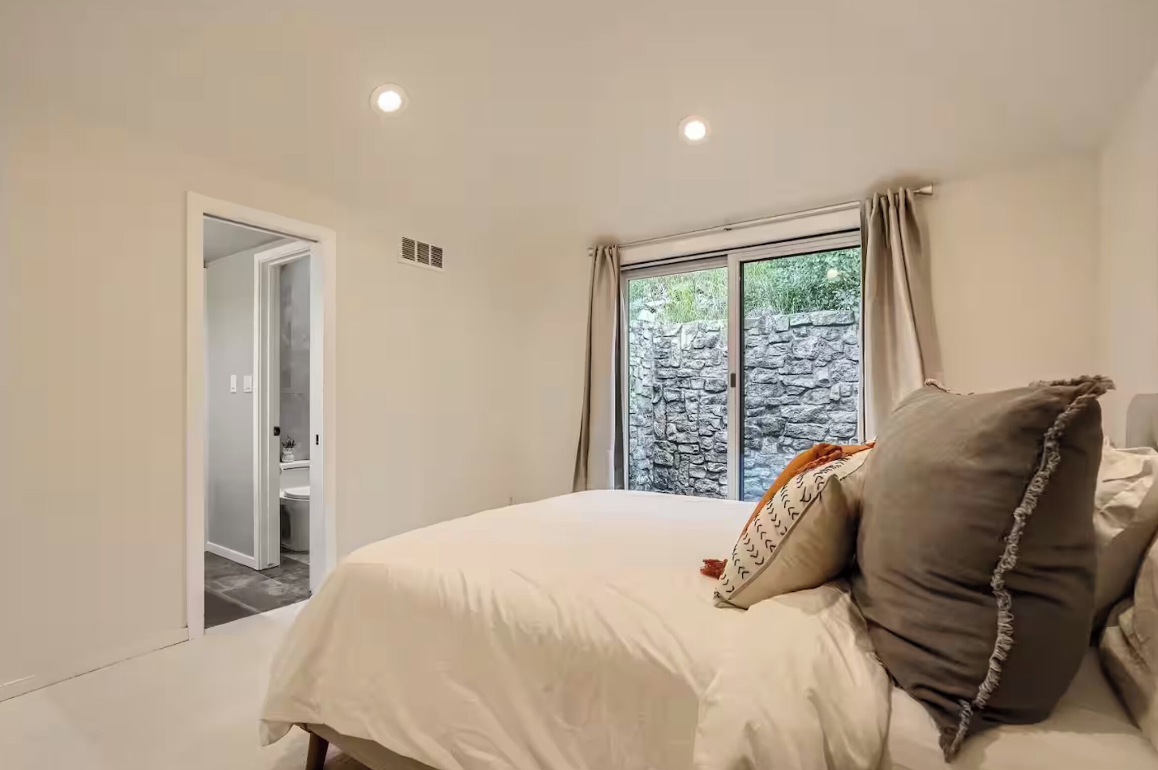 A bedroom with a bed, a window with curtains, and a view of a stone wall outside.
