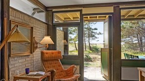Une pièce chaleureuse avec un mur en briques, une table en bois surmontée d’une lampe, un fauteuil à motifs écossais et une vue sur l’extérieur.