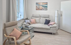 A modern living room with a glass coffee table, a beige sofa with multiple cushions, and a large window with curtains.