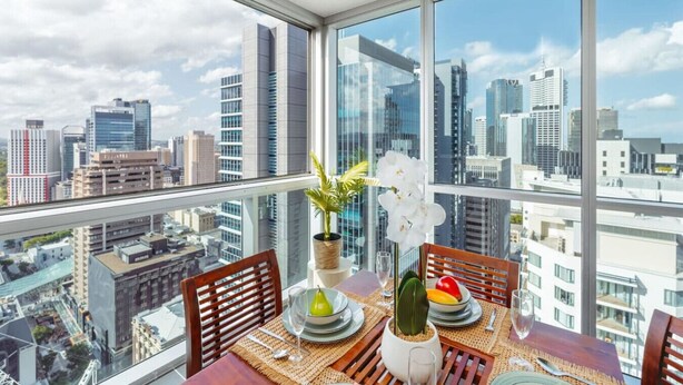 A dining area with a table set for two, a potted plant, and a view of a cityscape through large windows. - Great Value CBD 2 Beds Apt Free Carpark/ Pool /Gym (Brisbane City)