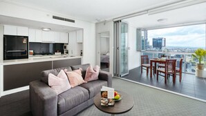 A modern apartment with a grey sofa, a dining area with wooden chairs, a kitchen with white cabinets, and a balcony with a city view. - Great Value CBD 2 Beds Apt Free Carpark/ Pool /Gym (Brisbane City)