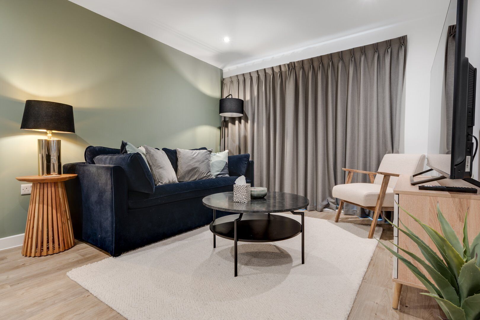 Un salon moderne avec un canapé bleu foncé, une table basse ronde, une table d’appoint en bois, une lampe, un fauteuil et un bureau avec un ordinateur.