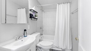 A modern bathroom with a white sink, toilet, and bathtub. There is a towel rack above the sink and a shower curtain. - Spacious Mid Century Modern 1 Bed 1 Bath 65" TV (Wyandotte)