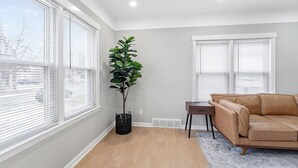 A modern living room with a beige leather sofa, a tall green plant in a black pot, and a small wooden table. - Spacious Mid Century Modern 1 Bed 1 Bath 65" TV (Wyandotte)
