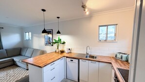 A compact kitchen with a wooden countertop, white cabinets, a built-in dishwasher, a sink, and a toaster. There is a small window above the sink and a potted plant on the countertop. - Cozy unit Papamoa Beach (Bay of Plenty)
