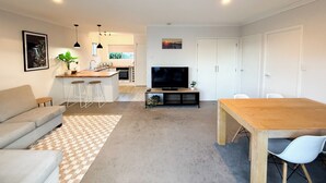 A modern living room with a sofa, a dining table, and a kitchen area with a TV. - Cozy unit Papamoa Beach (Bay of Plenty)