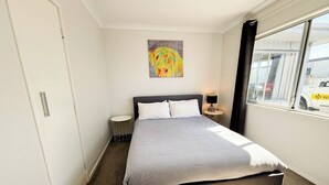 A hotel room with a large bed, a small bedside table, a wall-mounted painting, and a window with a view of a vehicle outside. - Cozy unit Papamoa Beach (Bay of Plenty)