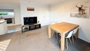 A modern kitchen with a built-in oven, a flat-screen TV on a wooden stand, and a dining area with a wooden table and white chairs. - Cozy unit Papamoa Beach (Bay of Plenty)