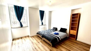 A bedroom with a wooden floor, a bed with patterned bedding, a wardrobe, and two windows with blue curtains.