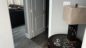 Een gang met een houten vloer, een deur naar een andere kamer en een klein tafeltje met een lamp.