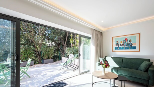 A modern living room with a green sofa, a small round table, and a glass door leading to an outdoor area with a patio and greenery.