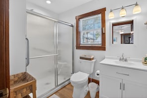 A bathroom with a glass-enclosed shower, a toilet, a sink with a mirror, and a window with blinds.