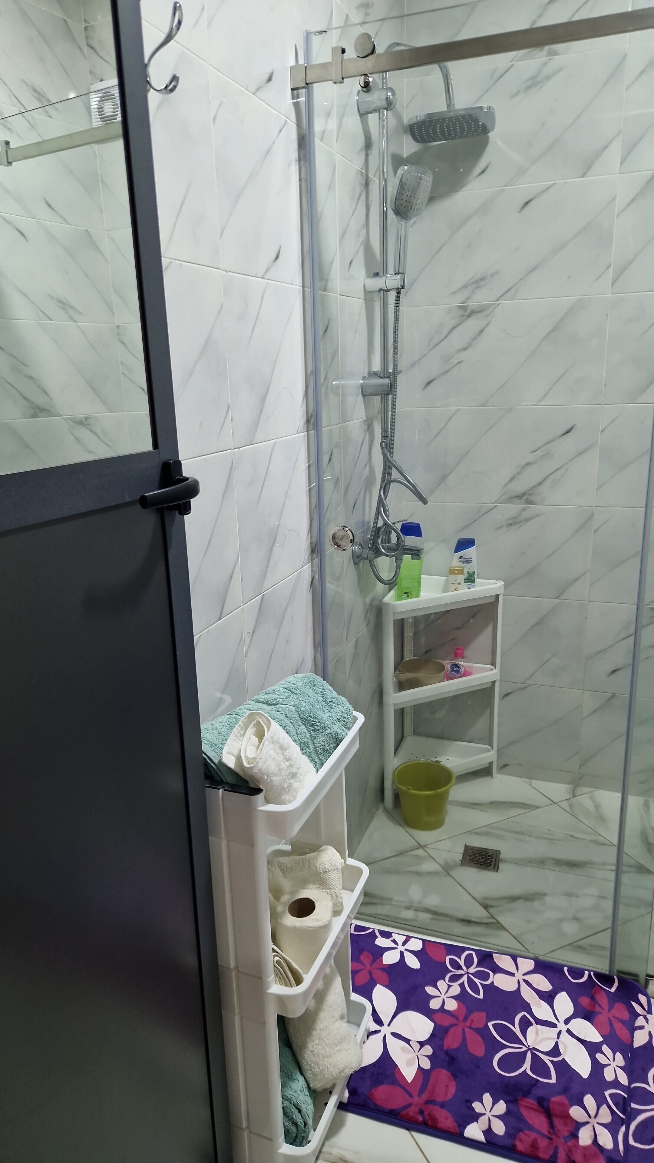 A shower area with a glass door, a shelf with toiletries, and a purple floral mat.
