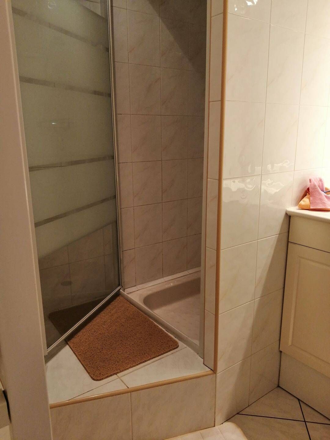 Une salle de bain d’angle avec une douche à parois de verre, un tapis de bain marron et un mur recouvert de carreaux blancs.
