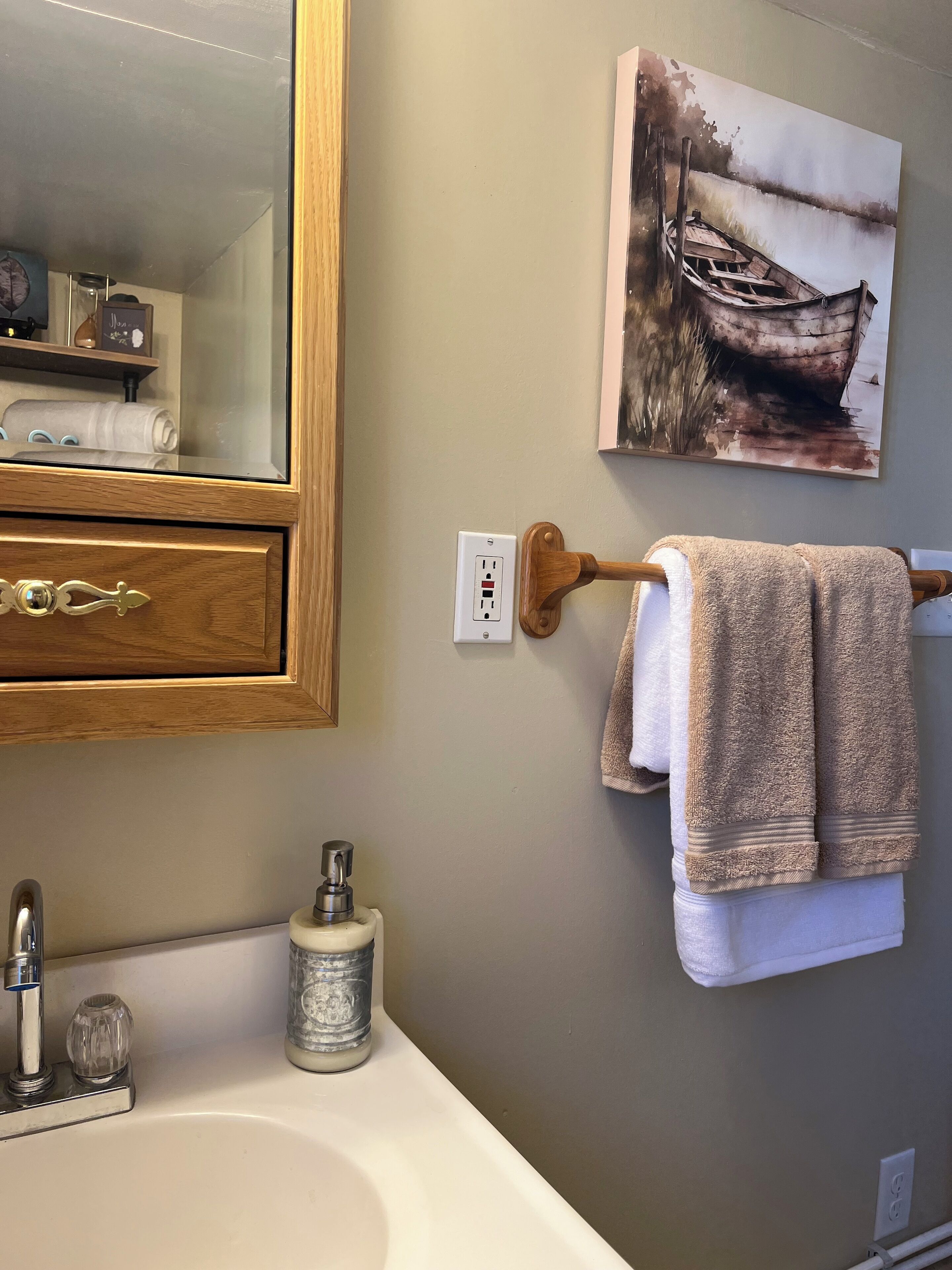 A bathroom with a wooden cabinet, a white sink, a mirror, a towel rack with towels, and a painting of a boat on the wall.