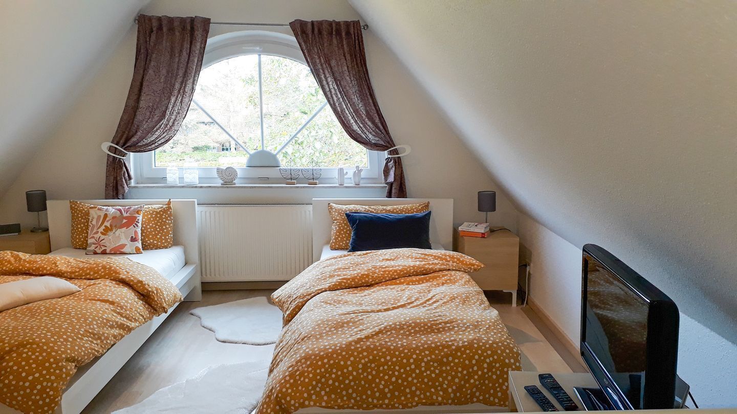 Un dormitorio con dos camas, una ventana con cortinas, una mesita de noche con lámpara y un televisor.