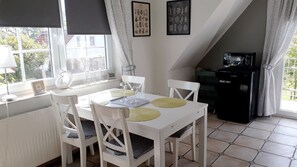 Comedor con una mesa y sillas blancas, un refrigerador negro y una ventana con cortinas.