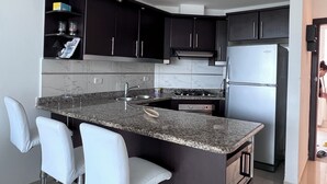 Une cuisine moderne avec des meubles de couleur sombre, un comptoir en granit, un évier en acier inoxydable et un four encastré. Trois chaises blanches sont disposées autour de l’îlot central.