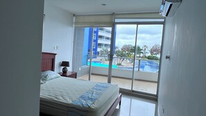 Une chambre d’hôtel avec un lit, une table de chevet, une lampe et une vue sur une piscine et les bâtiments environnants.