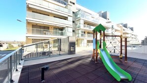 A playground with a slide and climbing structure on a rooftop terrace with a view of a modern building in the background.