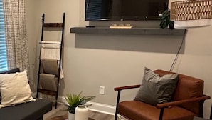 Una sala de estar con un sofá, un sillón de cuero marrón, una mesita redonda, una mesita auxiliar pequeña, una escalera, un televisor en un estante y una alfombra estampada.