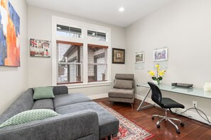 A modern living room with a grey sofa, a small armchair, a glass desk, and a chair. There are framed pictures on the wall and a window with blinds. - Luxury Lake Waterfront Friendly Retreat (Federal Way)