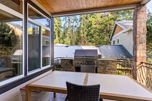 Un patio al aire libre con una mesa y sillas, una parrilla de piedra y vistas a una casa y árboles.