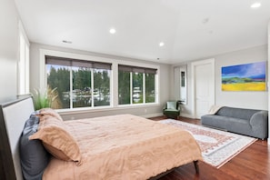 Un dormitorio con una cama grande, un sofá gris, una ventana con vistas a un lago y un cuadro en la pared.