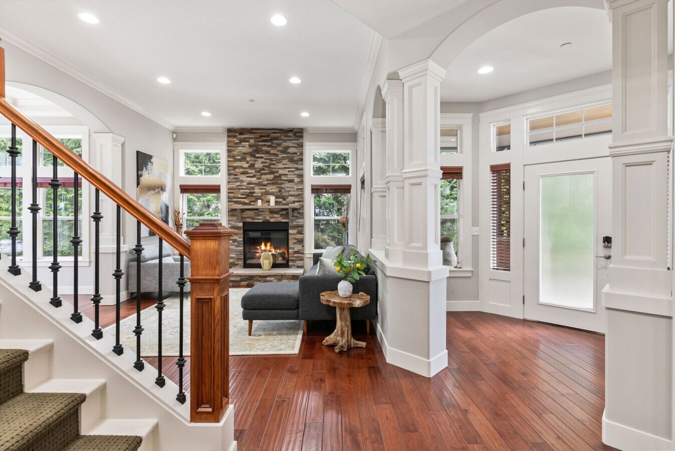 A spacious foyer with a wooden staircase, a stone fireplace, and a seating area with a small table and plant.