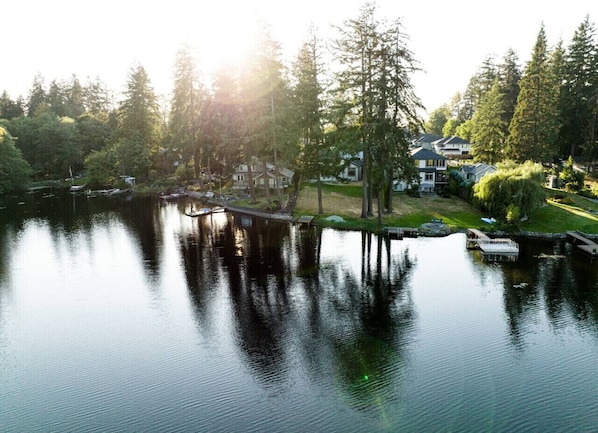 Una vista tranquila de un lago con un muelle de madera que se adentra en el agua, rodeado de abundante vegetación y edificios residenciales.